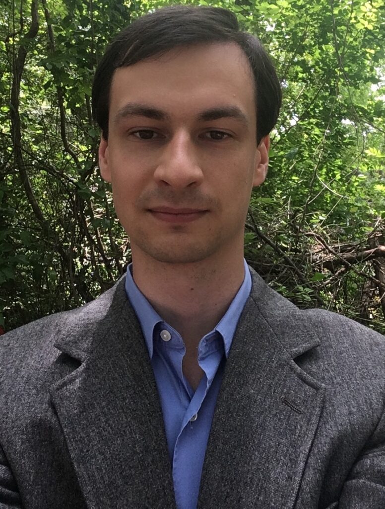 A man with brown hair gazes at the camera. He wears a pale blue button up and a grey jacket over it.