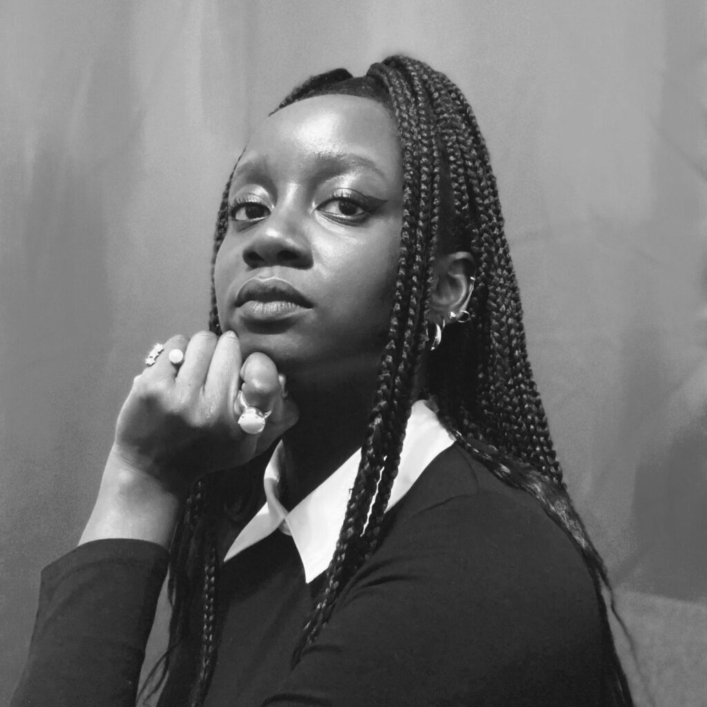 A woman with a long, braided ponytail gazes at the camera, resting her chin on her hand. She has several rings on her hand and several earrings along her left ear. The photo is in black and white.