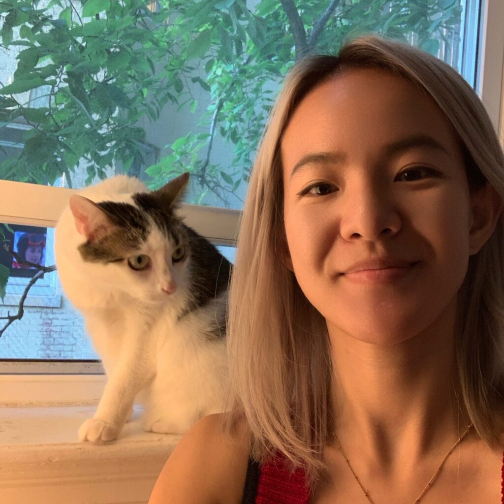A woman with dyed blonde hair smiles at the camera. She wears a gold necklace and a red tank top. A mostly white tabby cat eyes her in the background from their perch on the window sill.