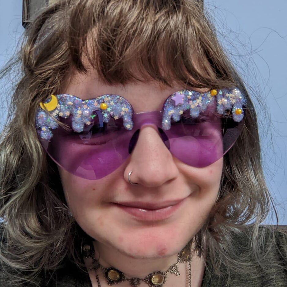 A woman with light brown hair and purple, heart-shaped sunglasses smiles at the camera.