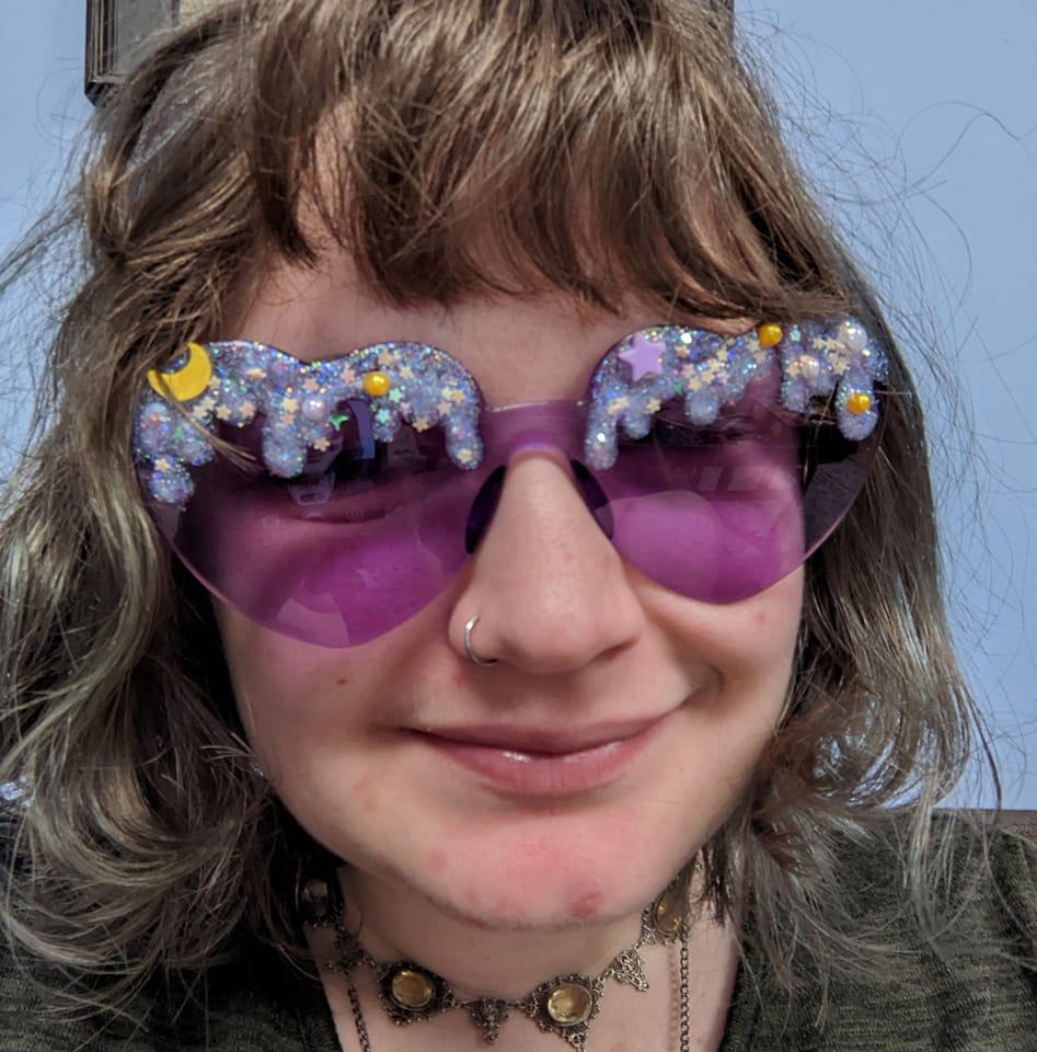 A woman with light brown hair and purple, heart-shaped sunglasses smiles at the camera.