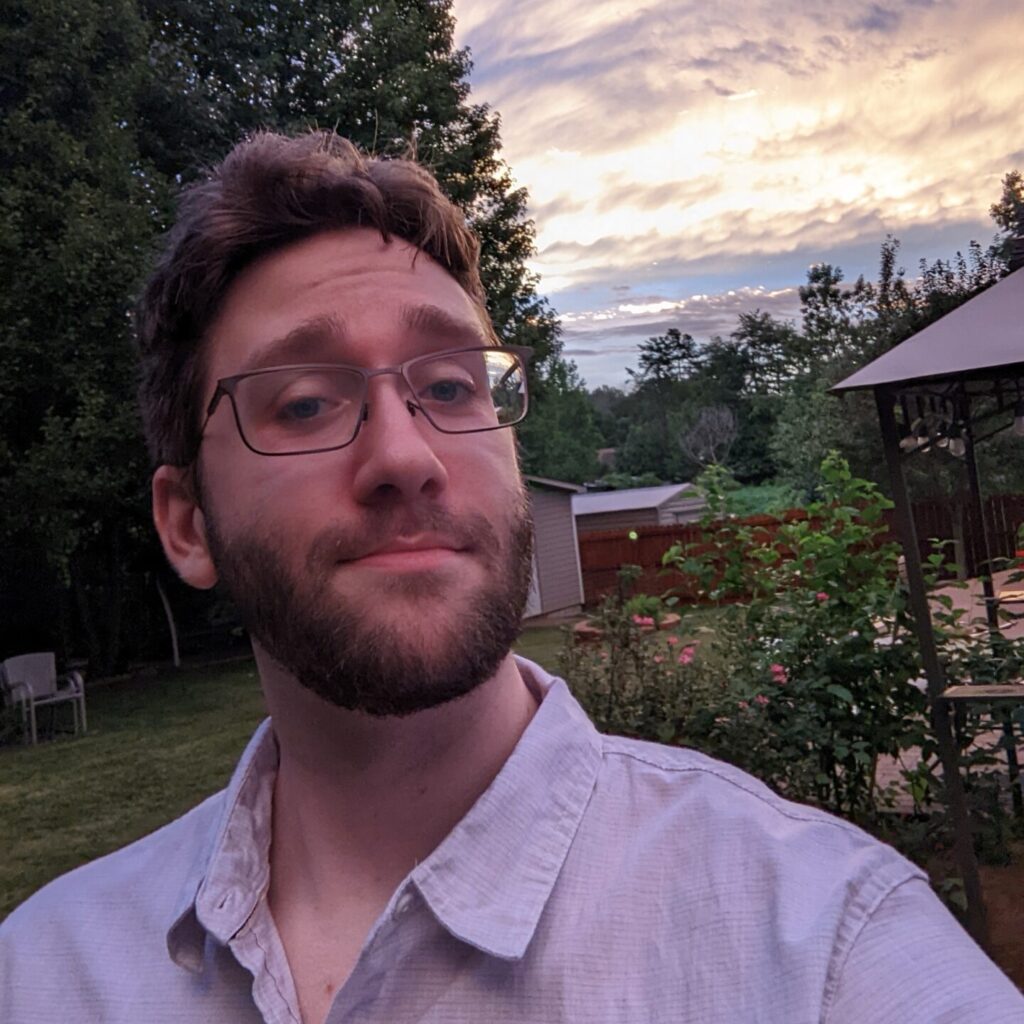 A man in a lavender button-up shirt and glasses smiles at the camera. He holds the camera selfie-style. He is outside and the background depicts clouds and pine trees.
