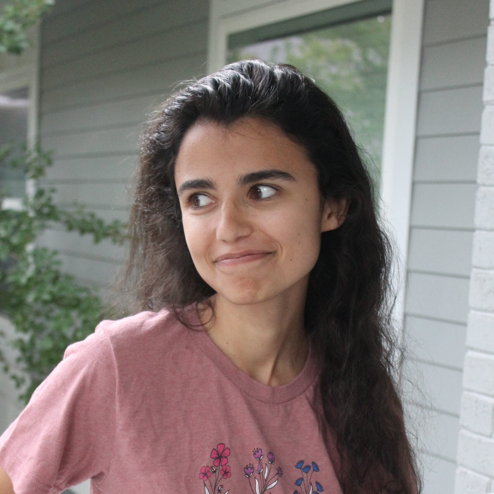 A person with long, dark hair smiles as she looks off to the left. She wears a pink shirt with pink, purple, and blue flowers on it.