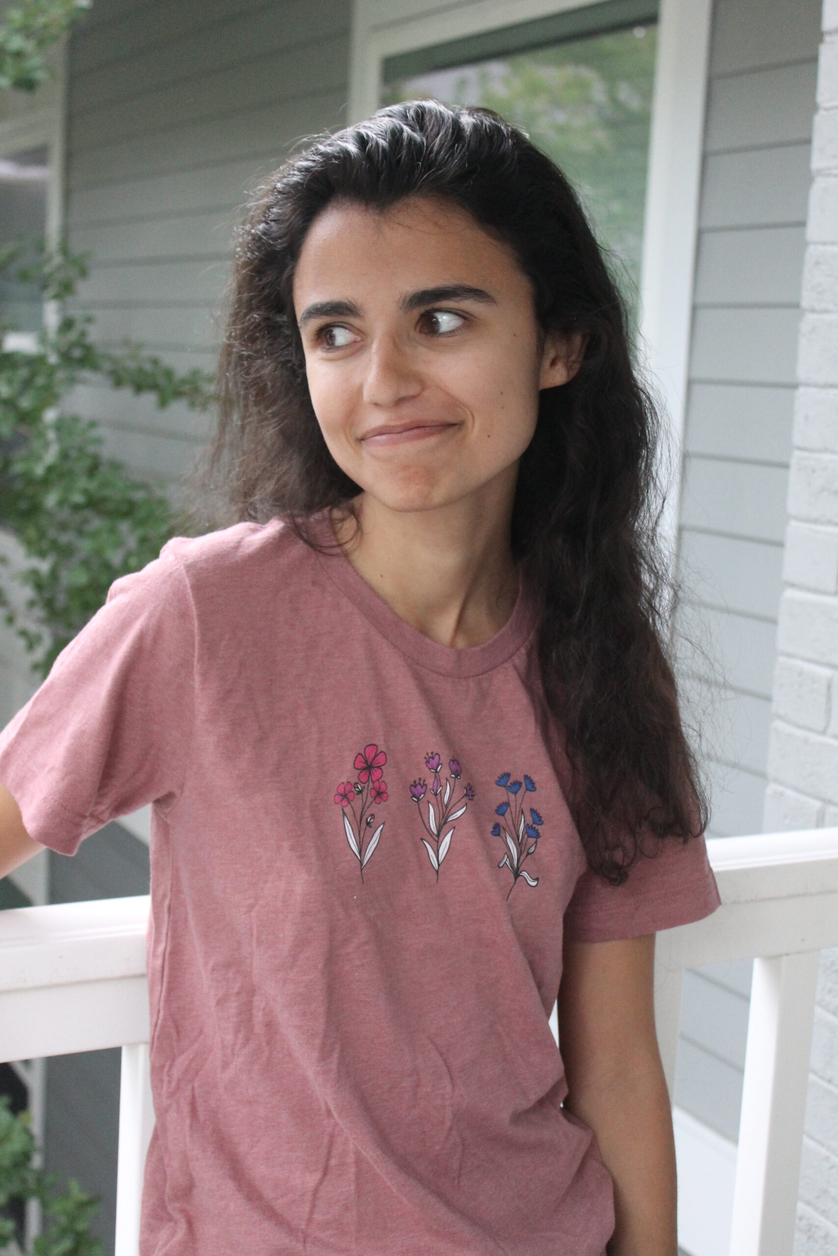 A person with long, dark hair smiles as she looks off to the left. She wears a pink shirt with pink, purple, and blue flowers on it.