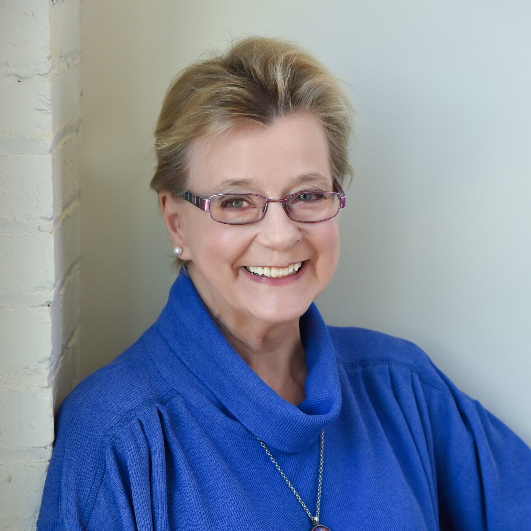 A woman with short, blonde hair and oval glasses smiles at the camera. She is wearing a dark blue sweater, pearl earrings, and a silver necklace. The background is white.