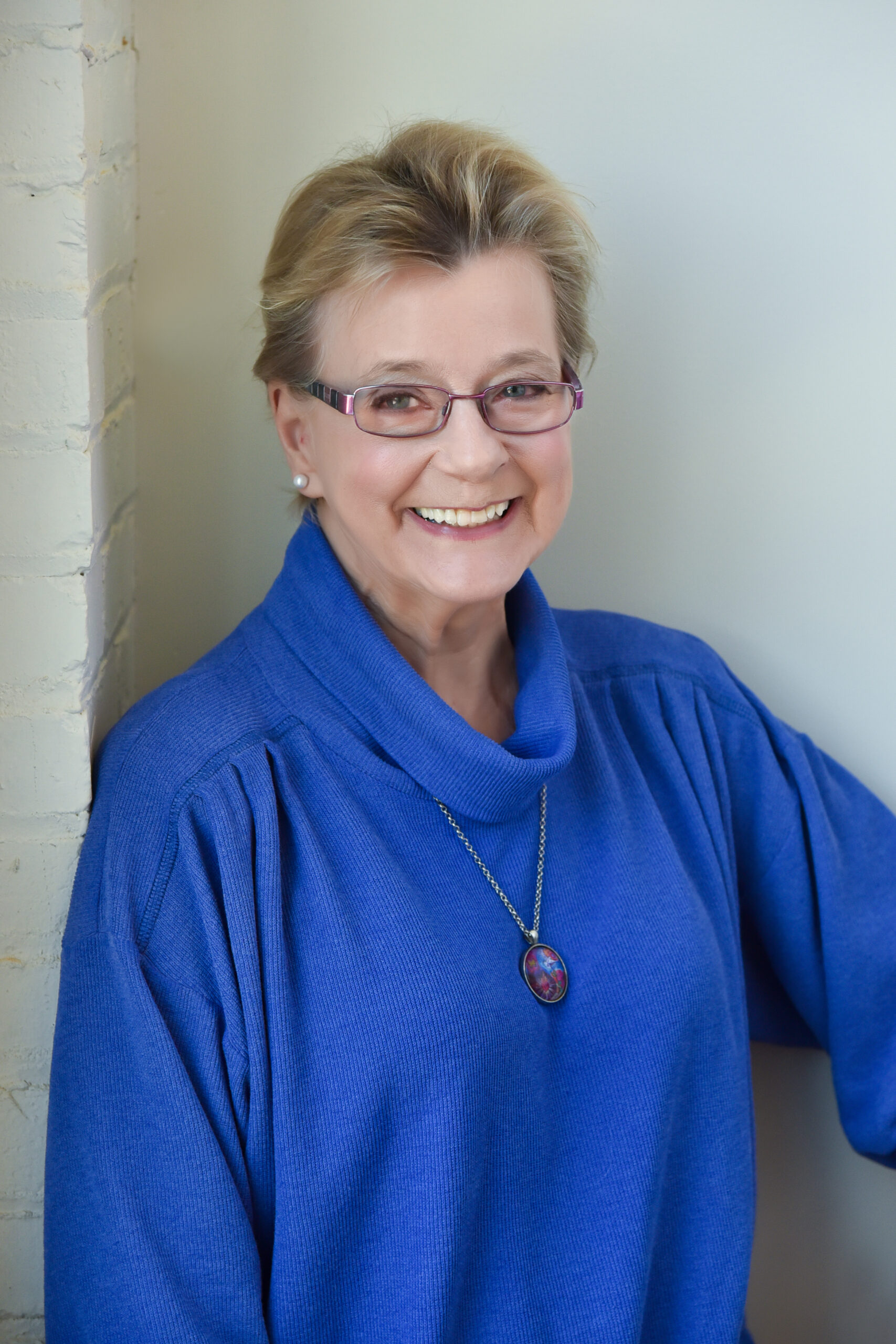A woman with short, blonde hair and oval glasses smiles at the camera. She is wearing a dark blue sweater, pearl earrings, and a silver necklace. The background is white.