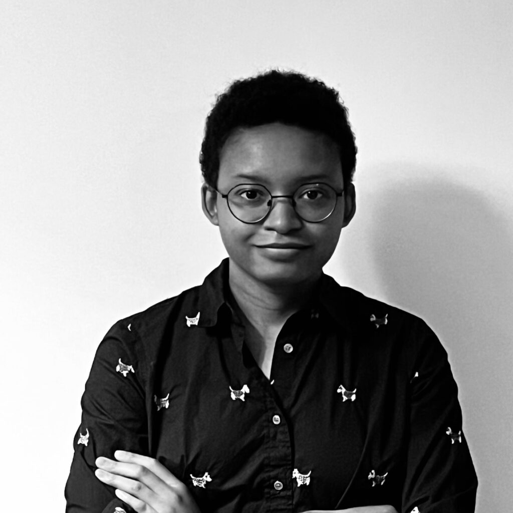 A black and white photo depicts a person with a small smile. They are crossing their arms over a black button up with small white dogs. Their hair is short and they wear round glasses.