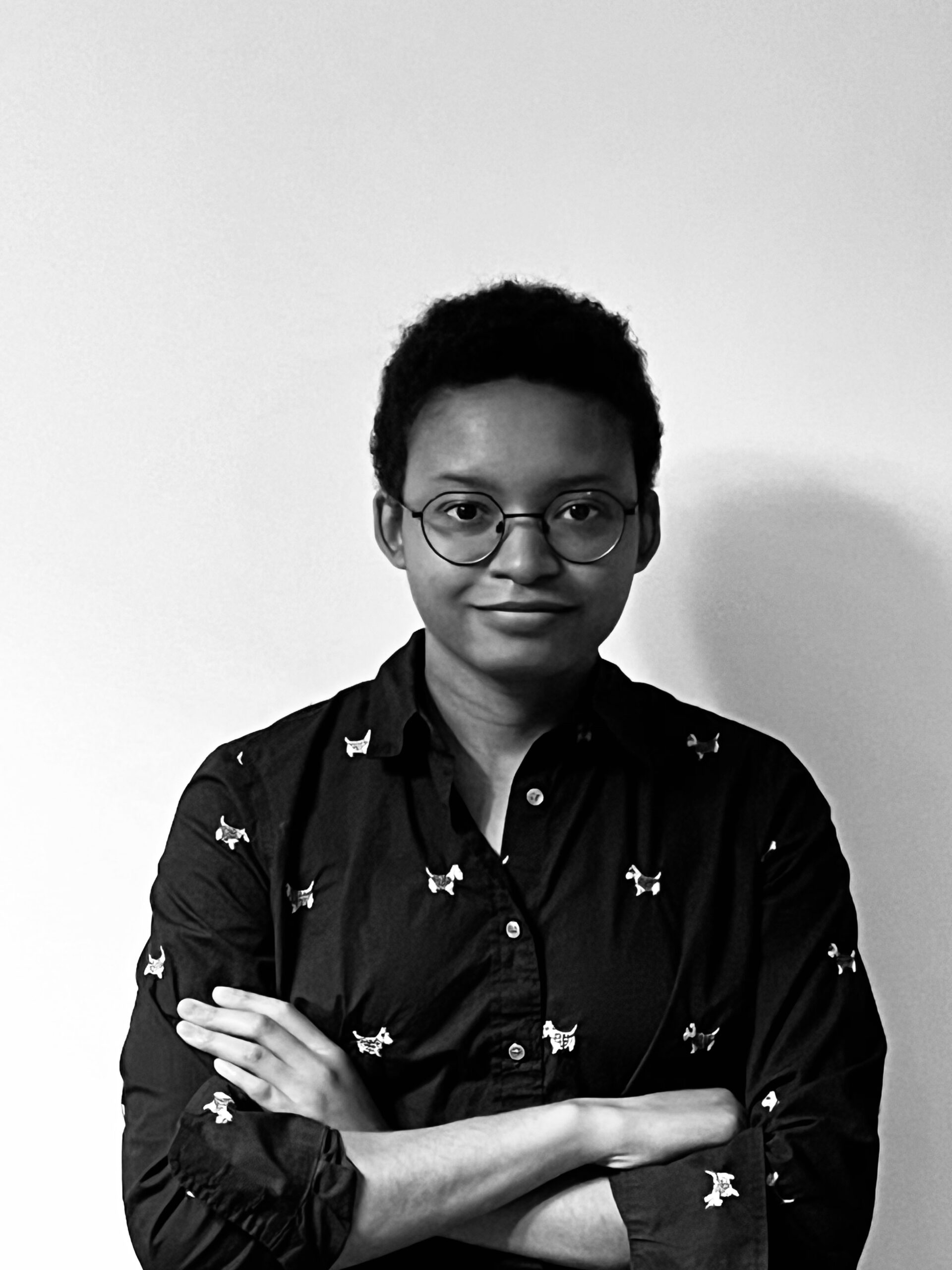 A black and white photo depicts a person with a small smile. They are crossing their arms over a black button up with small white dogs. Their hair is short and they wear round glasses.