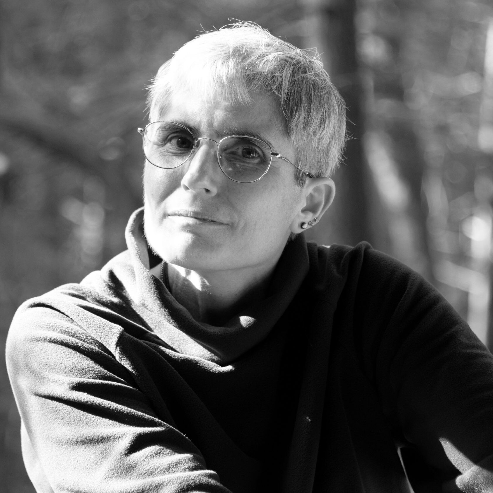 A white woman with short hair wears round glasses and a black sweater. She gazes at the camera. The photo is in black and white.