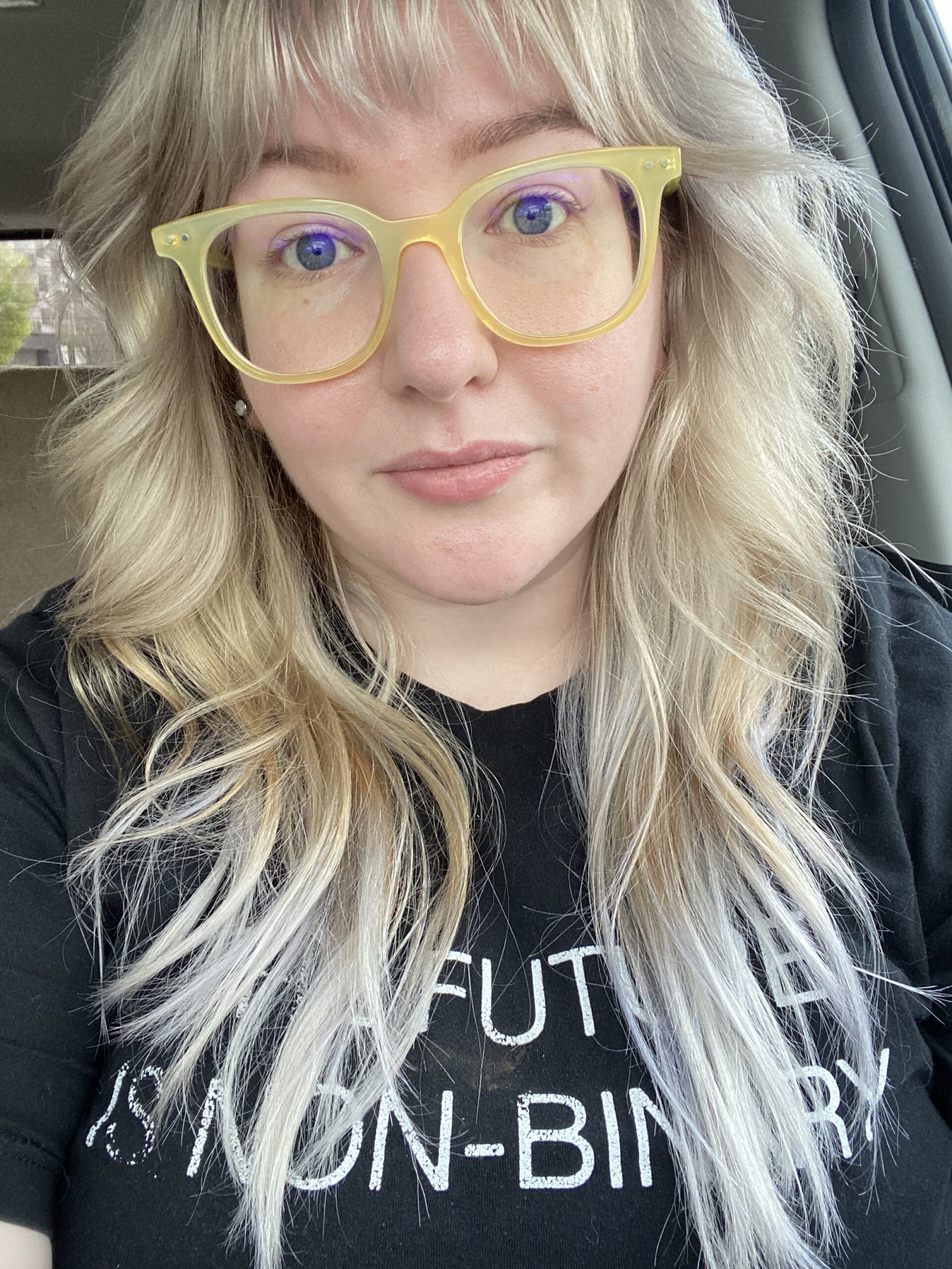 A white person with yellow-rimmed glasses and long, blonde hair. Their shirt reads, "THE FUTURE IS NONBINARY."