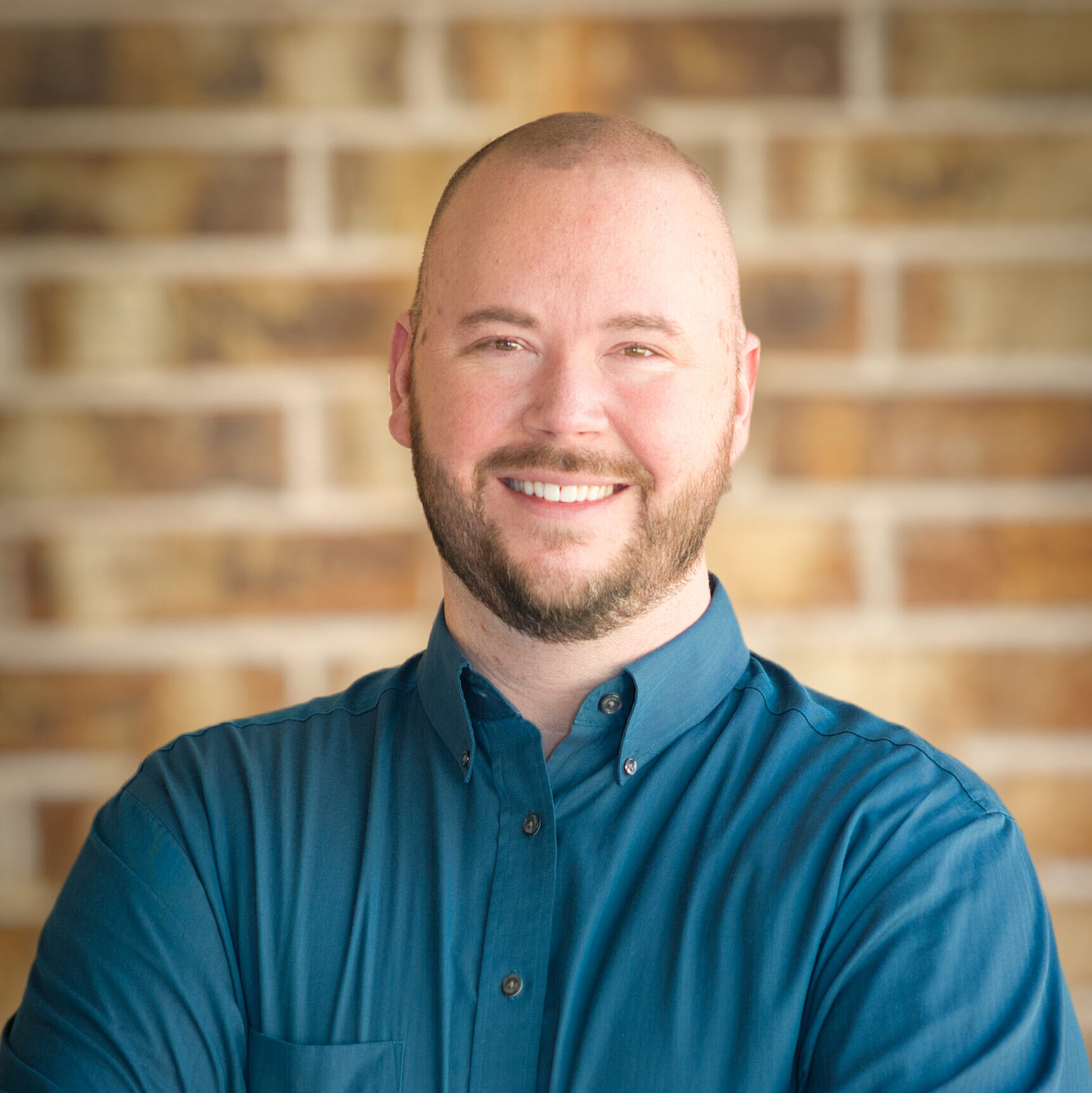 A bald man in a blue button up smiles at the camera. His arms are crossed. The background is blurred brick.
