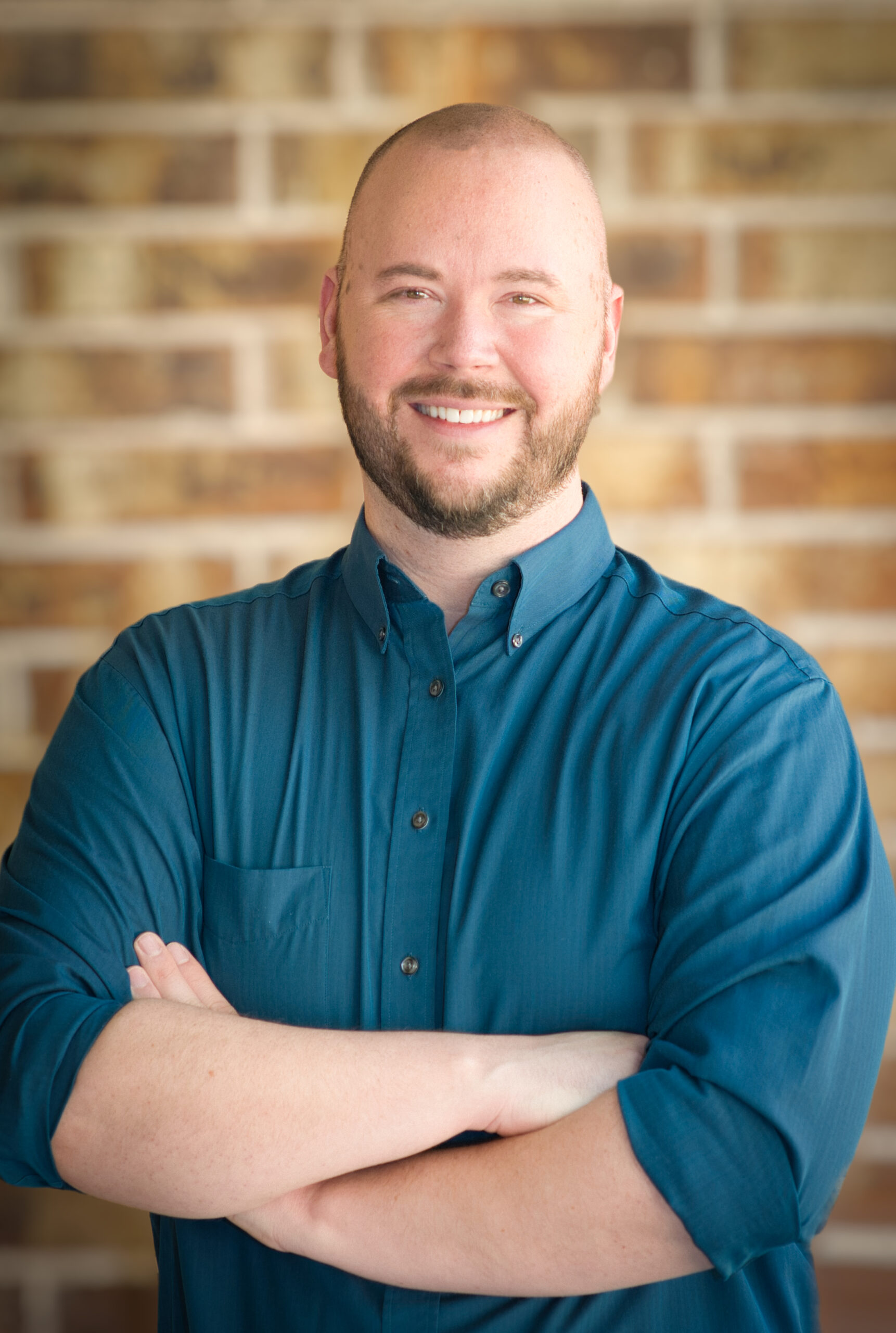 A bald man in a blue button up smiles at the camera. His arms are crossed. The background is blurred brick.