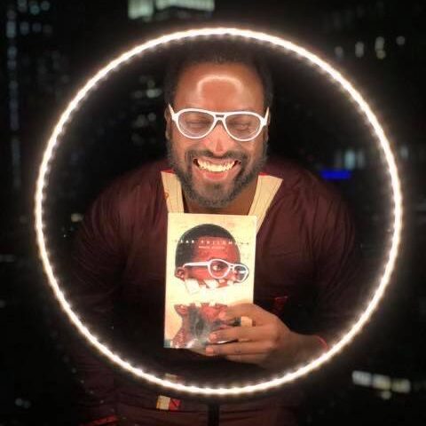 A Black person with white-rimmed glasses smiles widely with their eyes closed. They are holding their book, DEAR PHILOMENA by Mugabi Byenkya, and are surrounded by a circle of light.