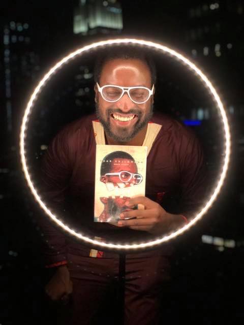 A Black person with white-rimmed glasses smiles widely with their eyes closed. They are holding their book, DEAR PHILOMENA by Mugabi Byenkya, and are surrounded by a circle of light.