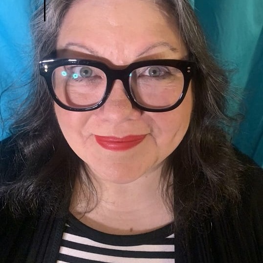 A woman with blue eyes, dark-rimmed glasses, and dark pink lipstick smiles with her mouth closed. Her hair is dark with gray in it and she wears a white and black striped shirt and a black jacket. The background is blue.