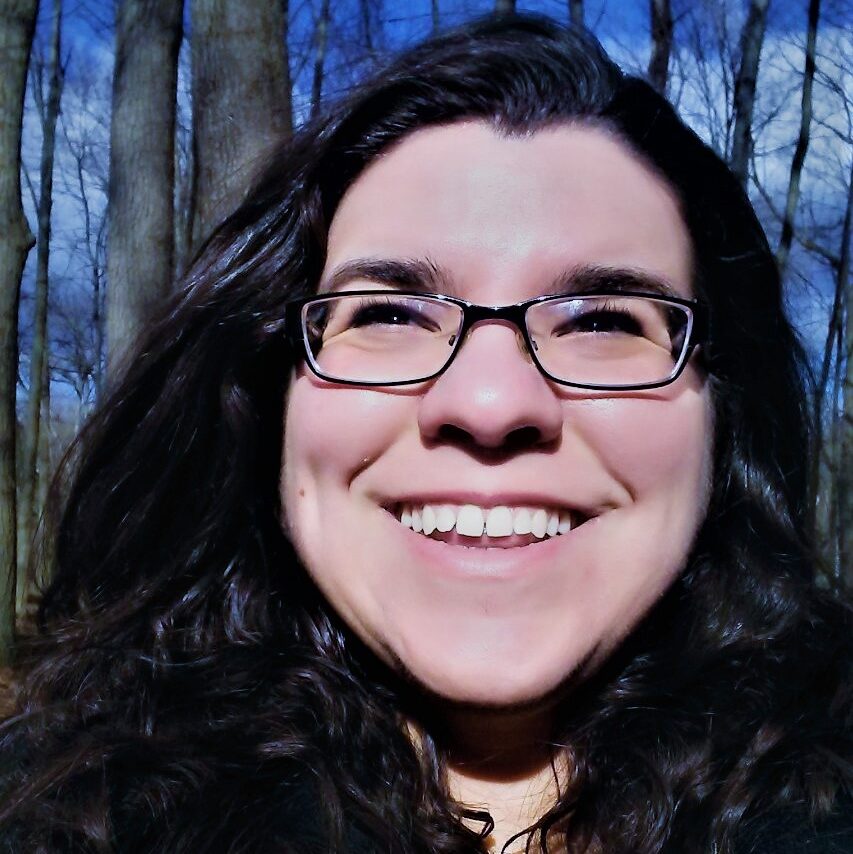 A white person, with long, dark hair and glasses smiles at the camera. The background is all thin threes and blue skies.