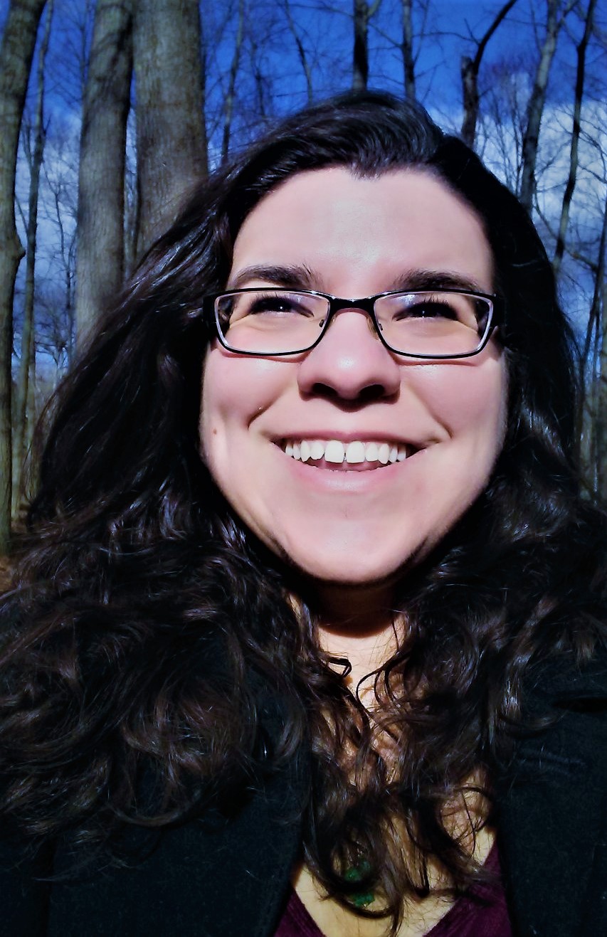 A white person, with long, dark hair and glasses smiles at the camera. The background is all thin threes and blue skies.