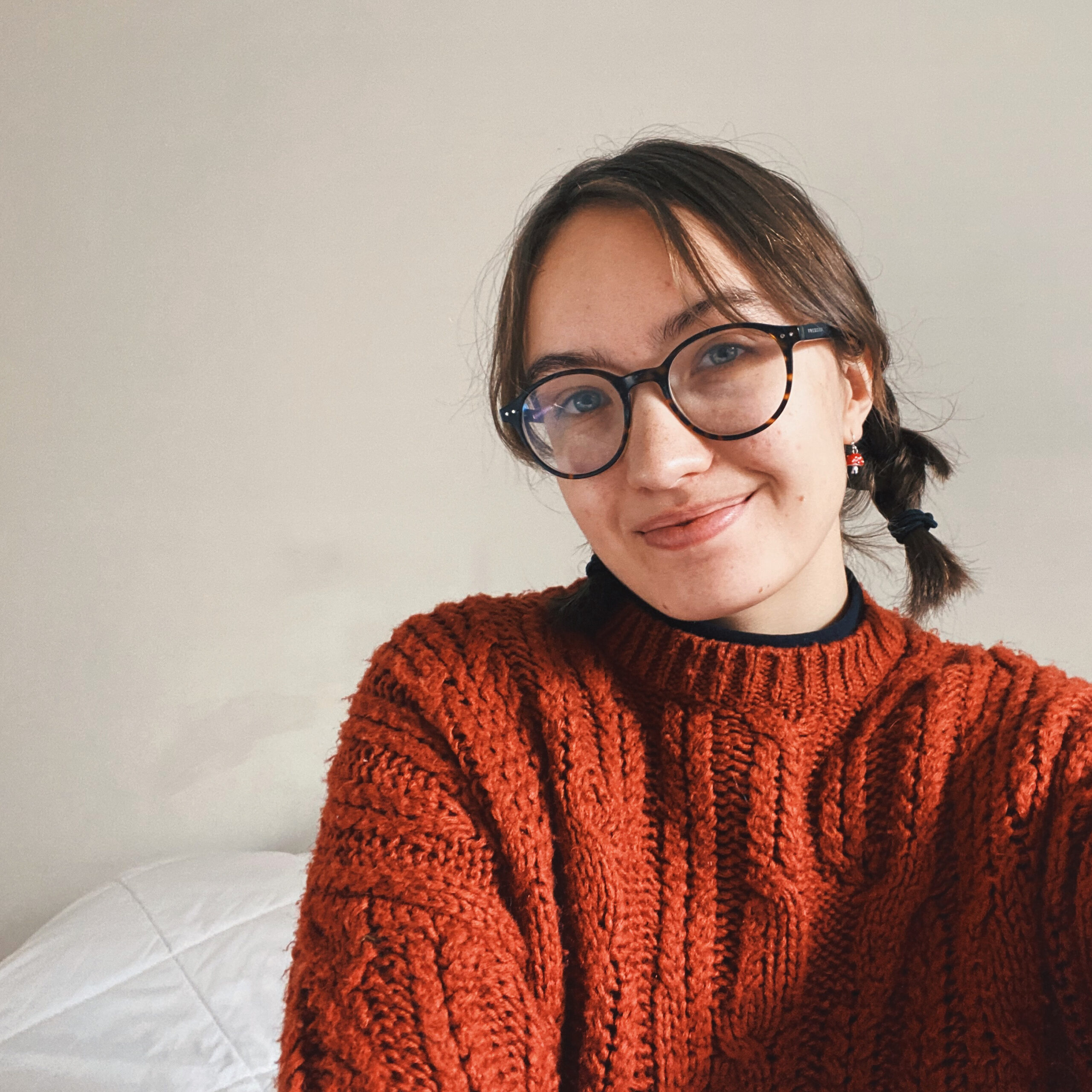 A white woman with short, brown hair in braids smiles at the camera with her mouth closed. She wears circular black glasses and an orange sweatshirt. The background is white.