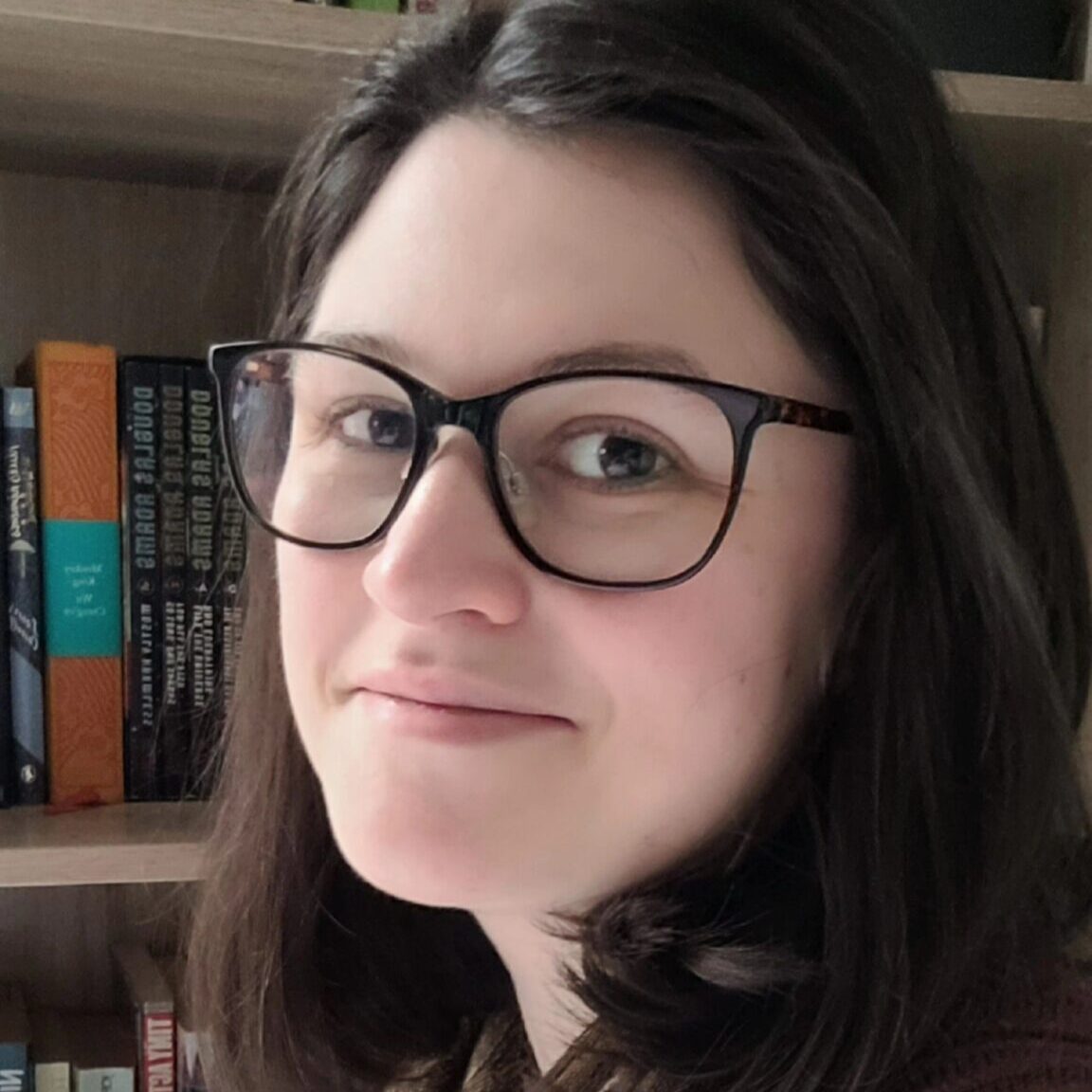 A white woman with dark hair and glasses gazes at the camera. Behind her is a shelf of books.