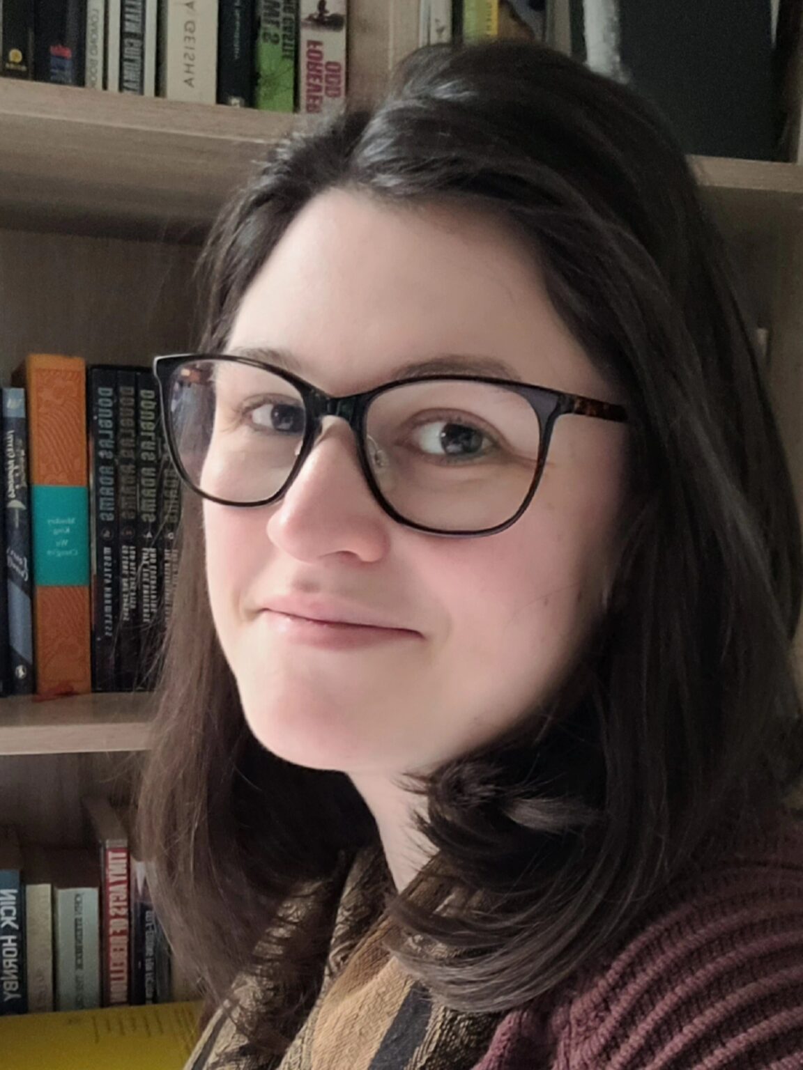 A white woman with dark hair and glasses gazes at the camera. Behind her is a shelf of books.
