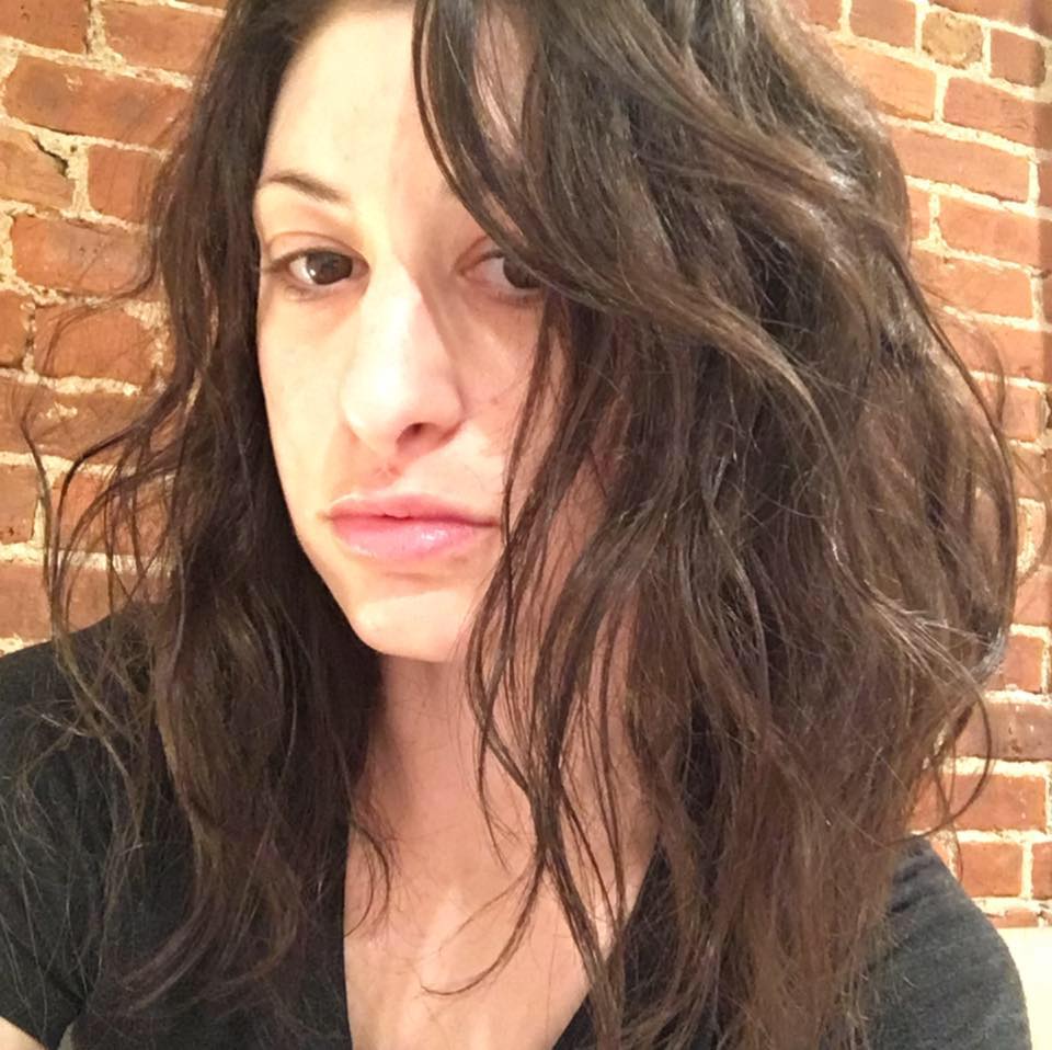 A white person with dark hair and brown eyes gazes at the camera. She wears a dark gray T-shirt. The background is red brick.