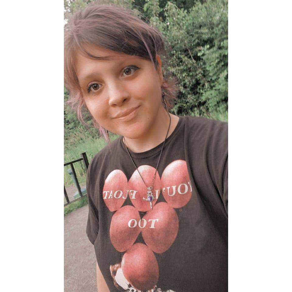 A white person with short, pink-tinted hair, smiling with her mouth closed at the camera. Her shirt reads, "You'll all float, too" and has the baloons on it from IT by Stephen King. The background is of pine trees.