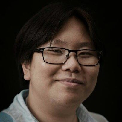 An Asian person with black-rimmed, rectangular glasses and short black hair against a black background. They wear a blue vest and are smiling with their mouth closed.
