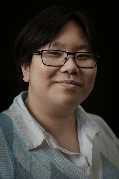 An Asian person with black-rimmed, rectangular glasses and short black hair against a black background. They wear a blue vest and are smiling with their mouth closed.