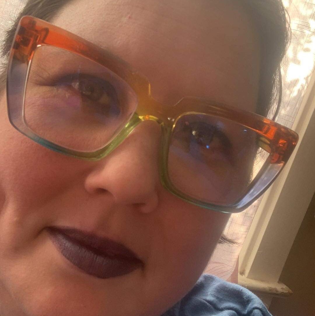 A white person with brown hair, colorful glasses, and purple lipstick wearing a blue shirt.