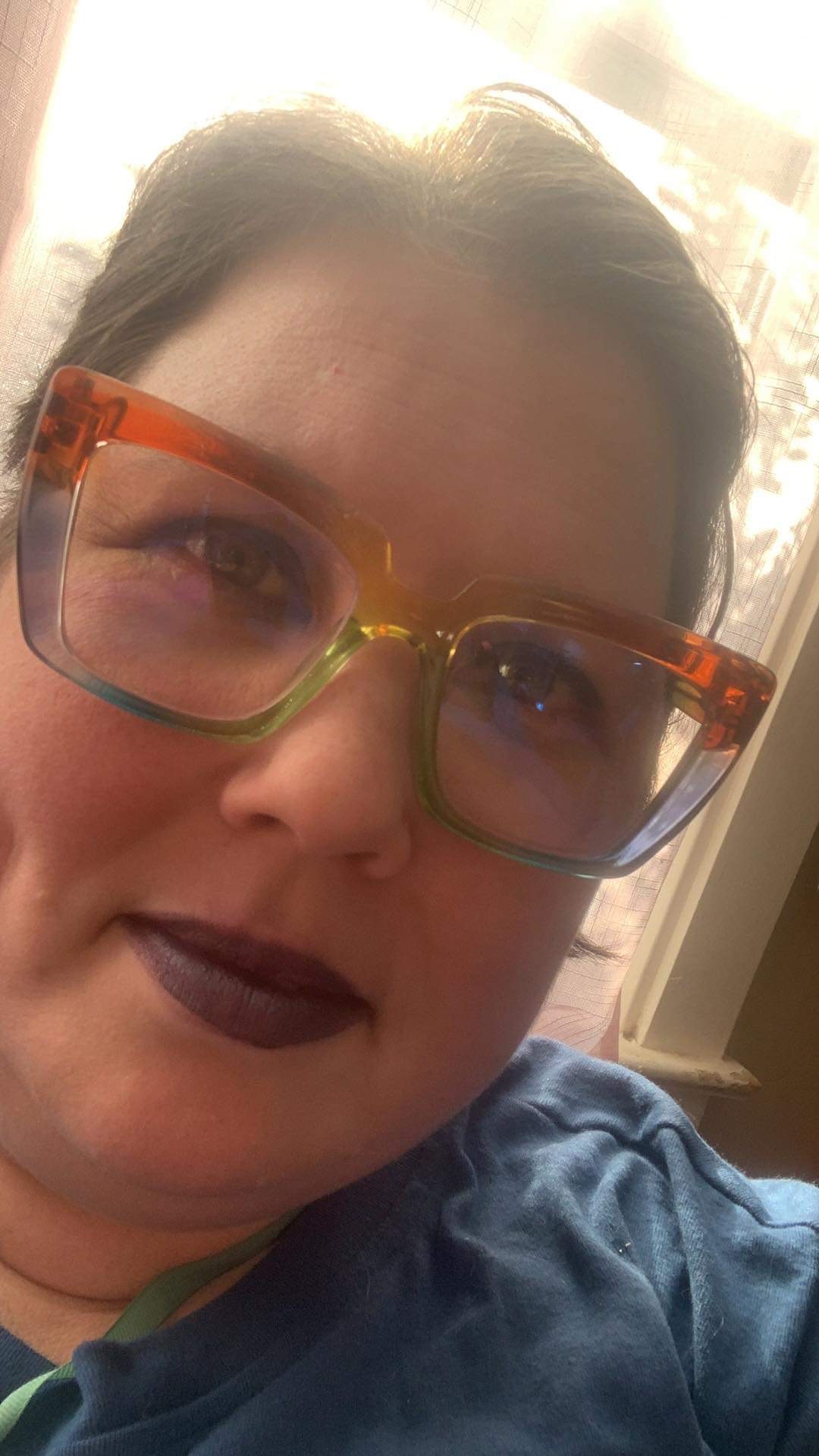 A white person with brown hair, colorful glasses, and purple lipstick wearing a blue shirt.