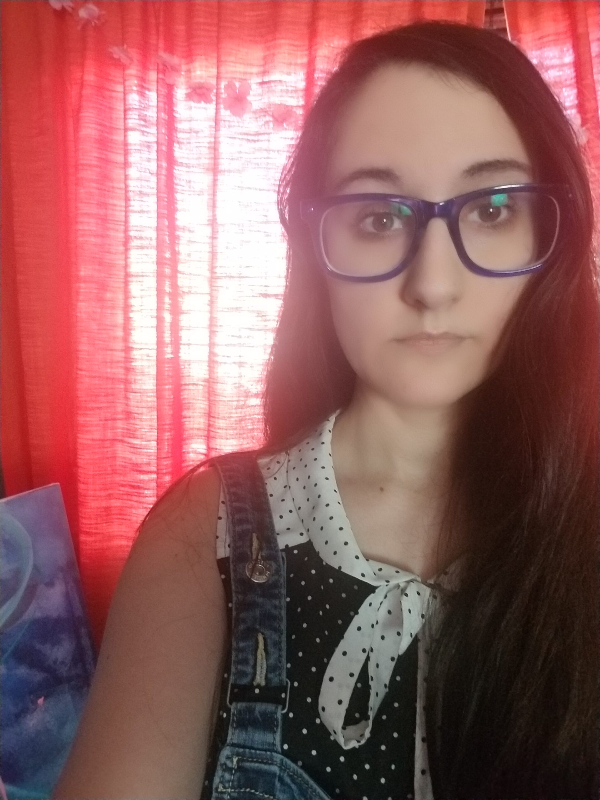 A white woman with long, dark hair wearing purple-rimmed glasses and a black and white spotted blouse takes a selfie in front of a transluscent red curtain.