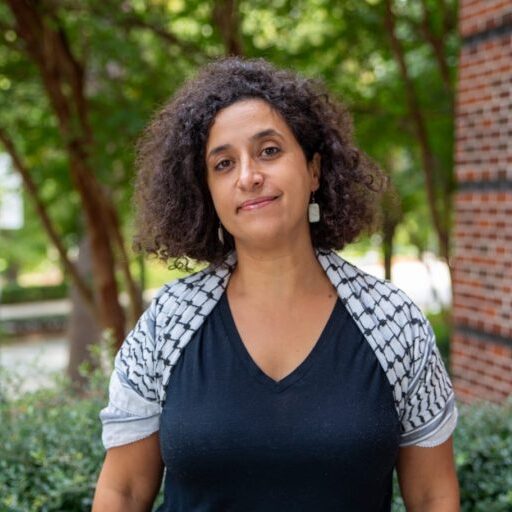 A woman with brown skin and shoulder-length black, curly hair wearing a blue shirt under a white and blue checked quarter-sleeve sweater. She is smiling at the camera with her mouth closed. The background is of trees and a brick building, all blurred.