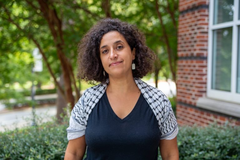 A woman with brown skin and shoulder-length black, curly hair wearing a blue shirt under a white and blue checked quarter-sleeve sweater. She is smiling at the camera with her mouth closed. The background is of trees and a brick building, all blurred.
