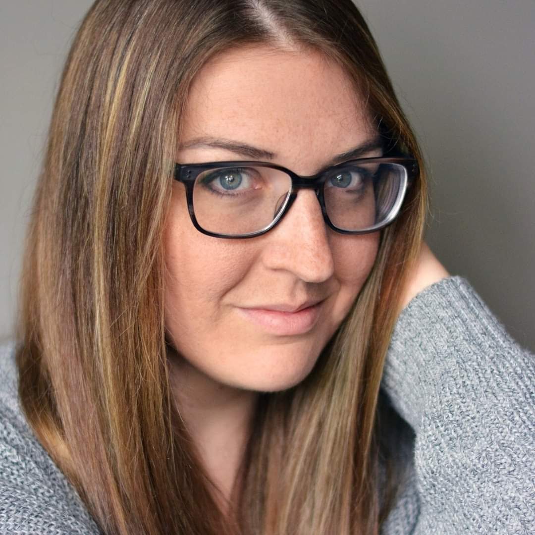 A white woman with straight, light brown hair and black-rimmed glasses. She is smiling at the camera with her mouth closed. Her sweater is light gray and the background is dark gray.