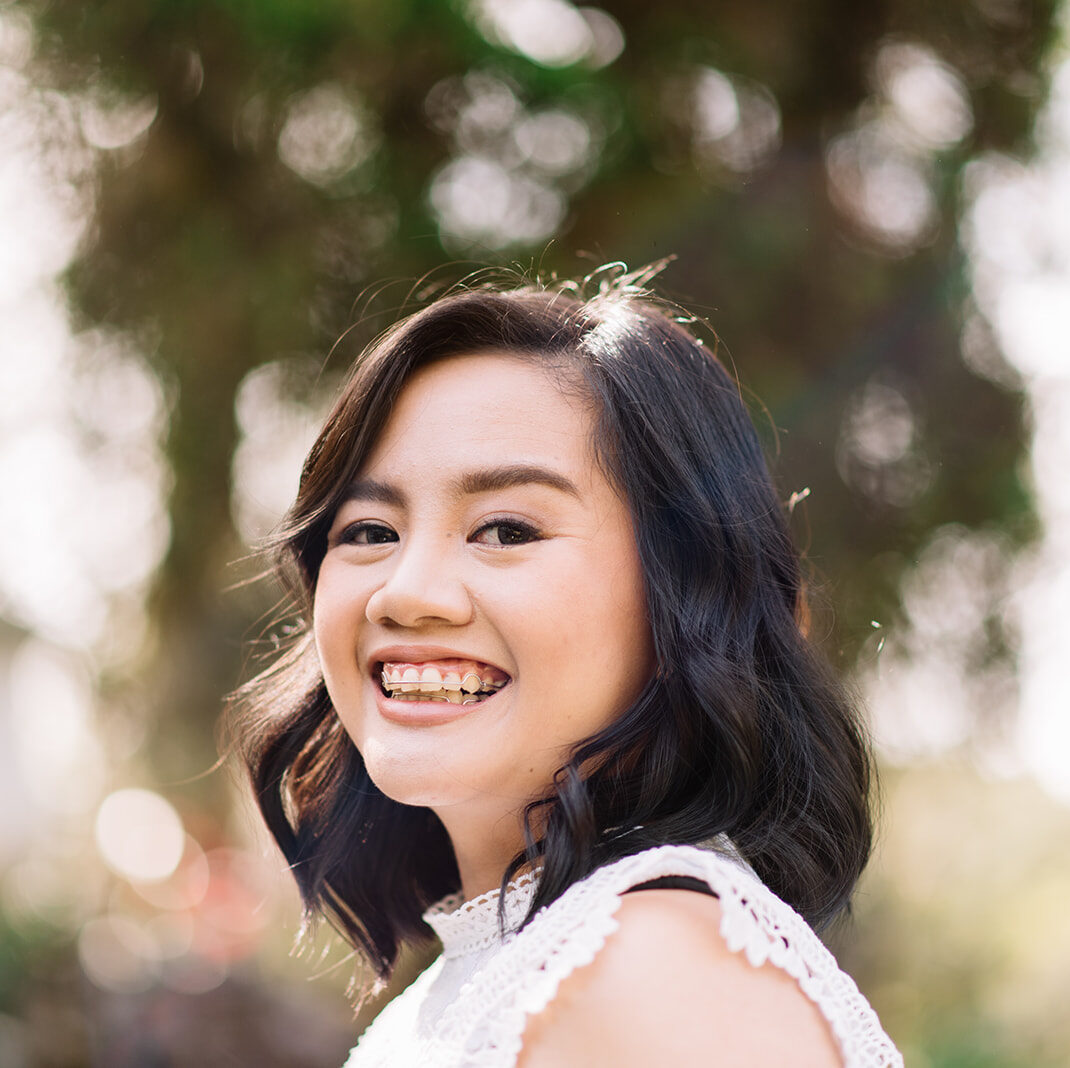 An Asian woman with black, shoulder length hair smiles widely at the camera. The background is blurred and seems to be trees.