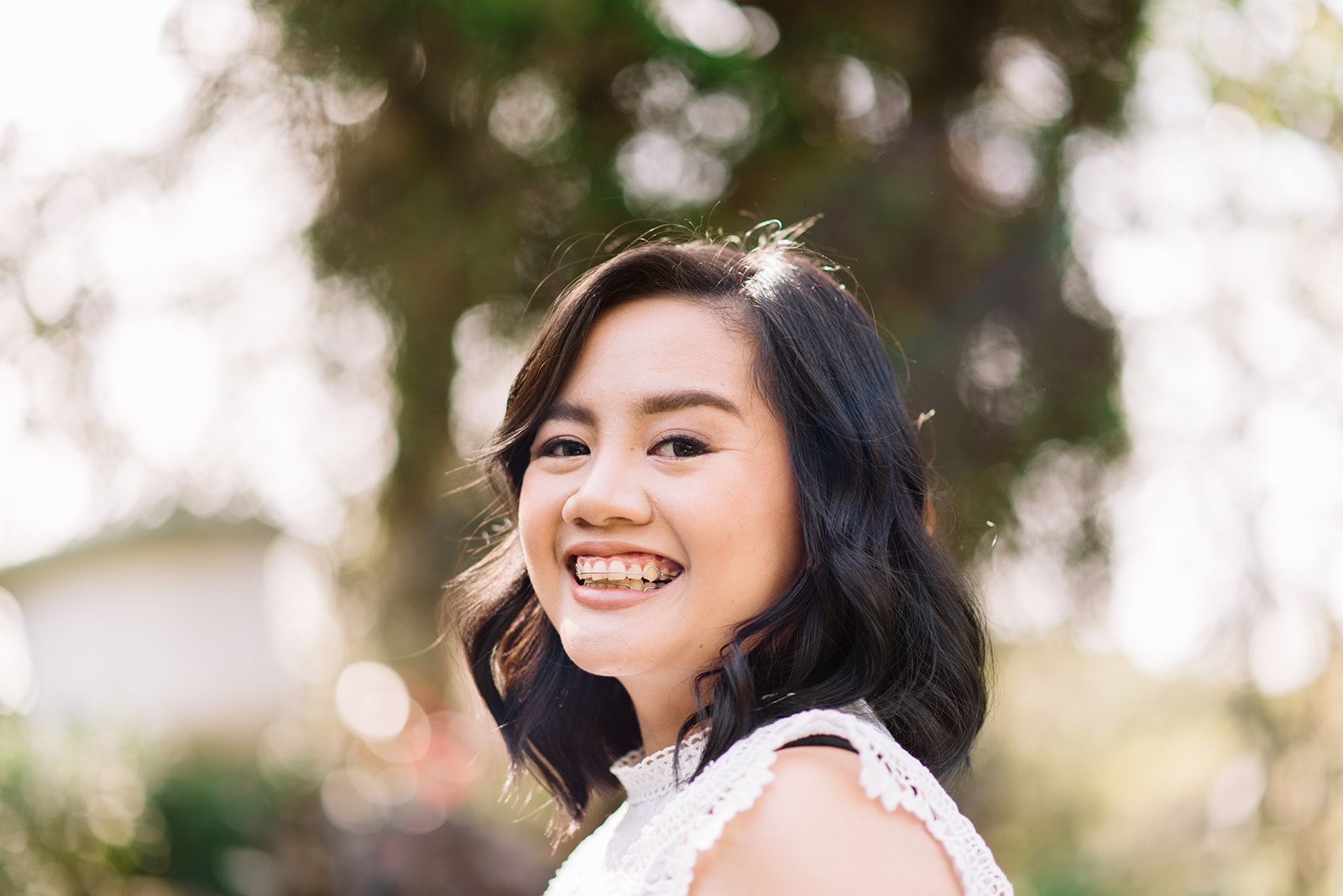 An Asian woman with black, shoulder length hair smiles widely at the camera. The background is blurred and seems to be trees.