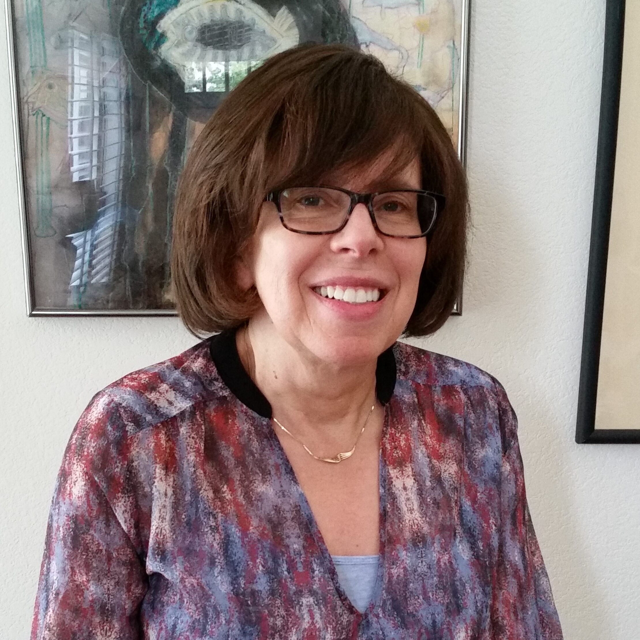 A white woman with a brown bob smiles at the camera. She wears black, square-rimmed glasses and a gold necklace.