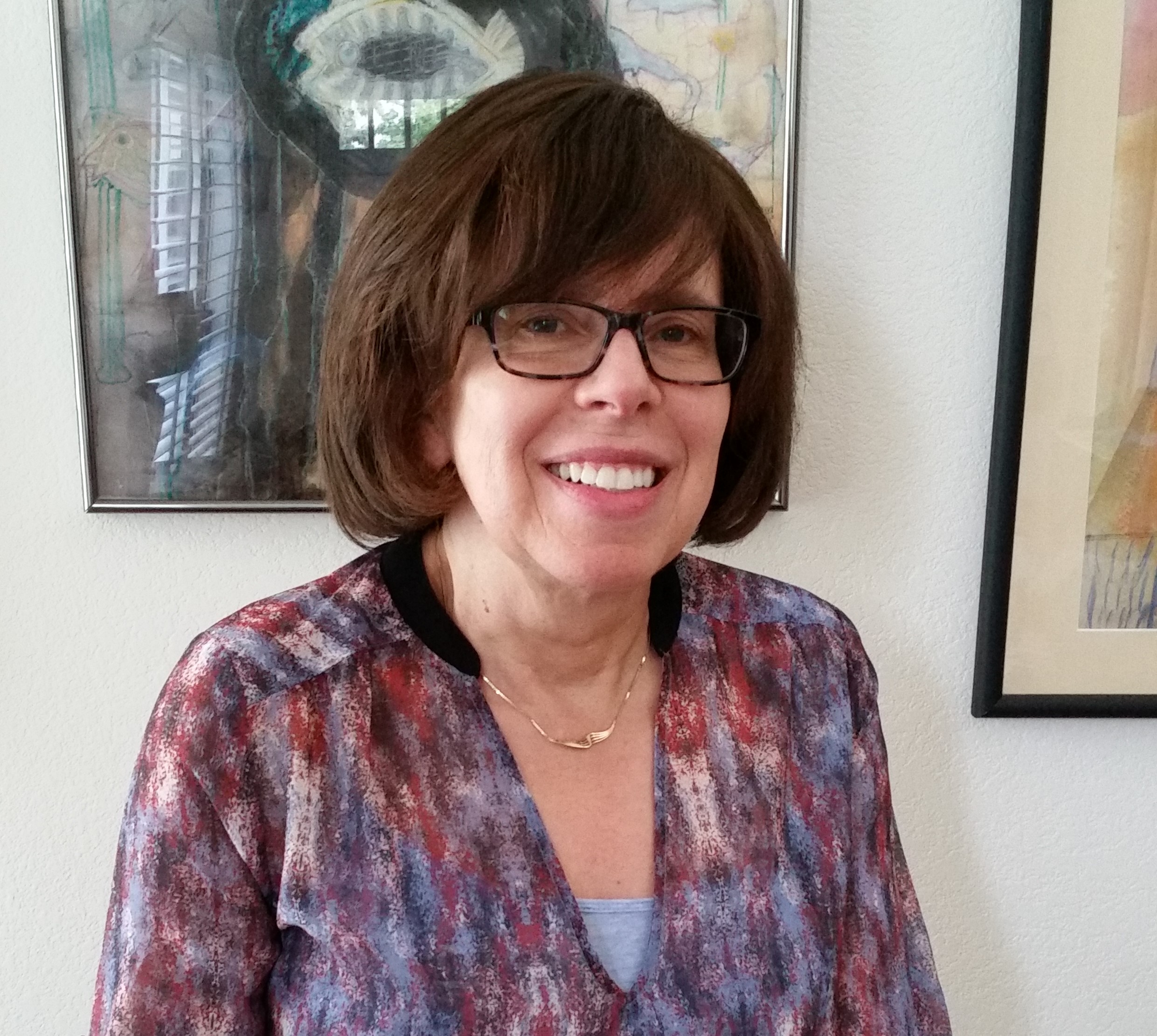 A white woman with a brown bob smiles at the camera. She wears black, square-rimmed glasses and a gold necklace.