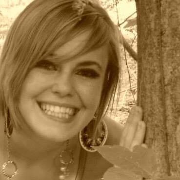A sepia-toned photo of a person with short, spiky hair wearing large, circular earrings and a pearly necklace. They are smiling and leaning against a tree.