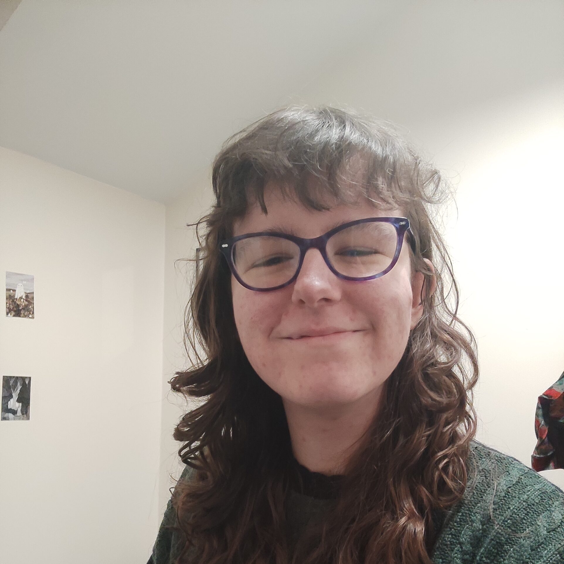 A white person with long, curly, brown hair wears a forest green sweater and black-rimmed glasses. She is smiling with her mouth and eyes closed.
