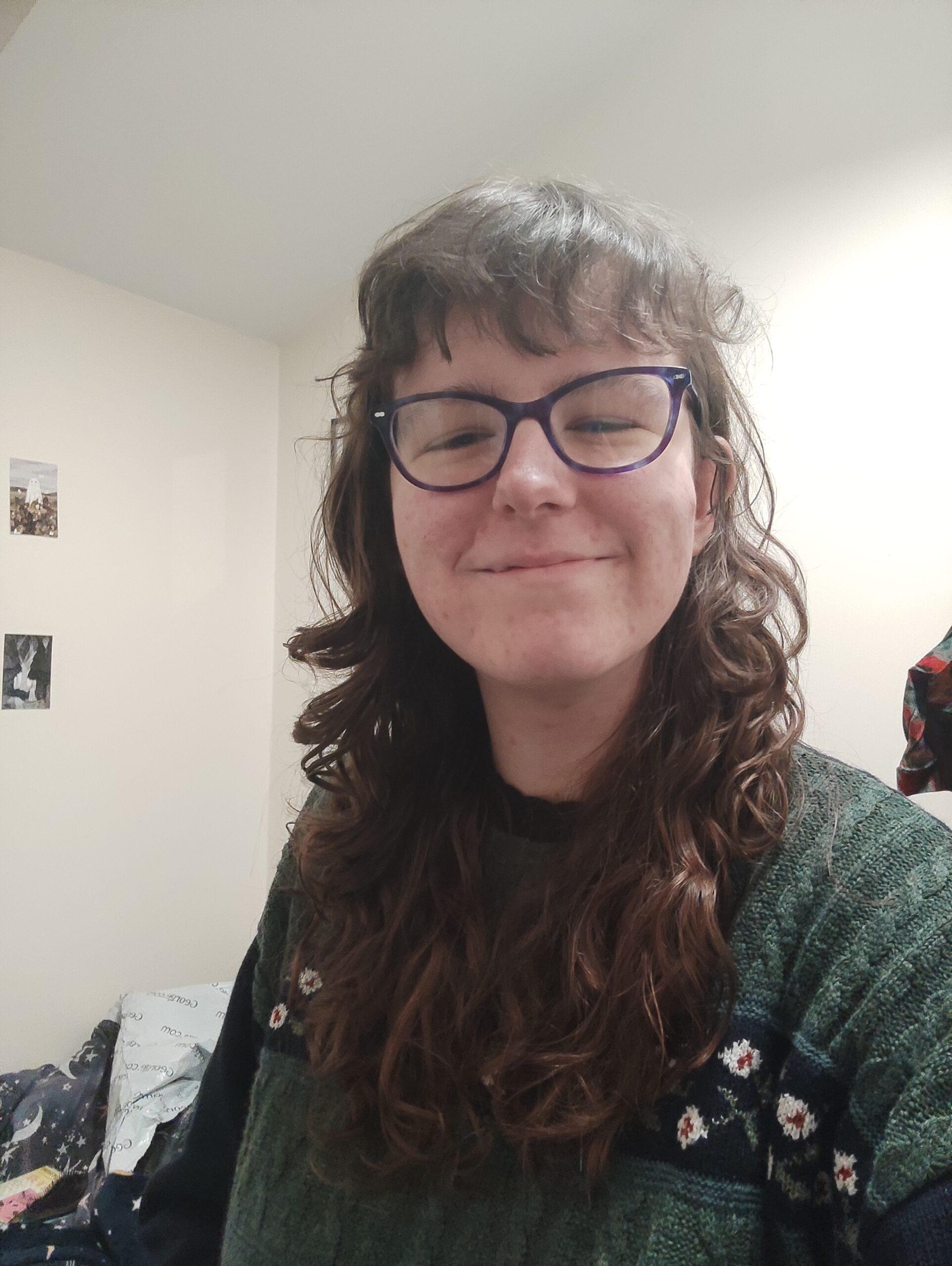 A white person with long, curly, brown hair wears a forest green sweater and black-rimmed glasses. She is smiling with her mouth and eyes closed.