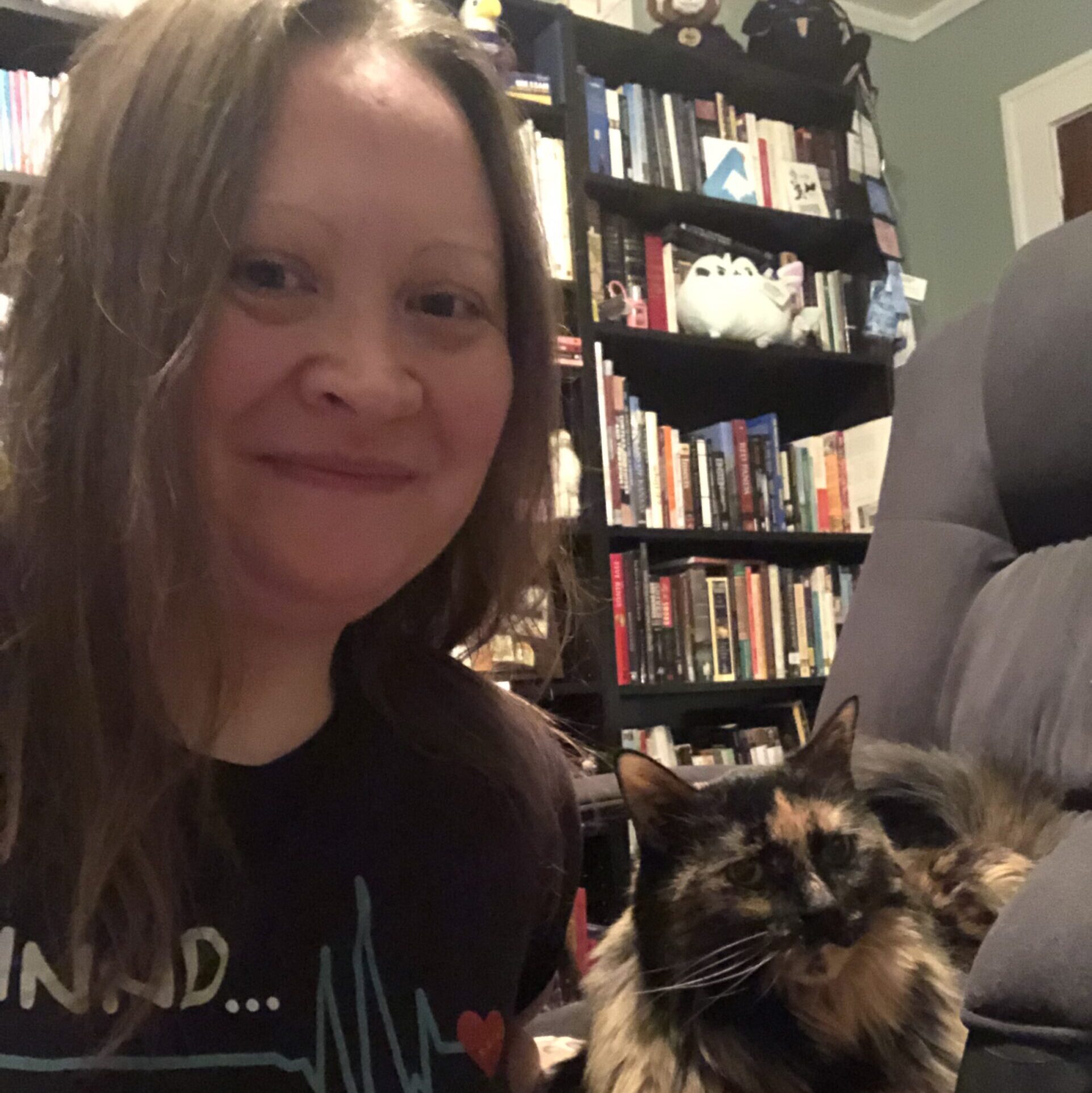 A white woman with light brown hair smiling with her mouth closed. The photo is a selfie. Also pictured: a bookcase in the background and a (really cute) brown and black cat.