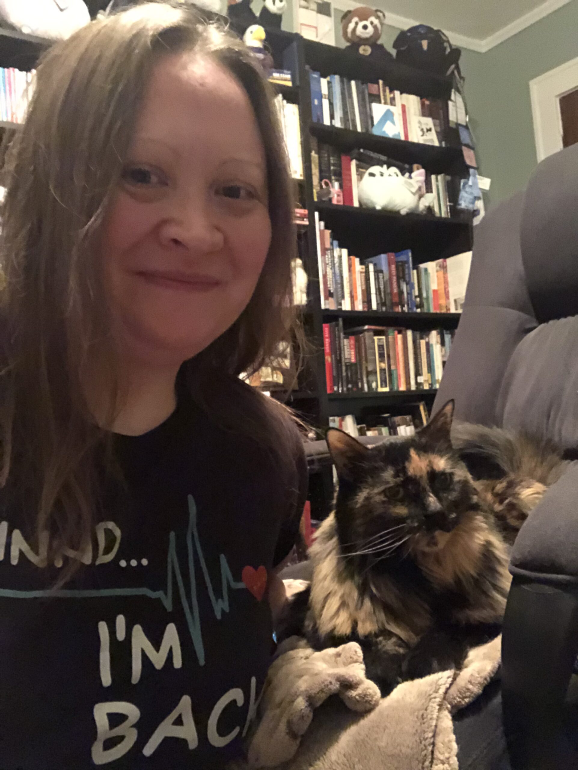 A white woman with light brown hair smiling with her mouth closed. The photo is a selfie. Also pictured: a bookcase in the background and a (really cute) brown and black cat.
