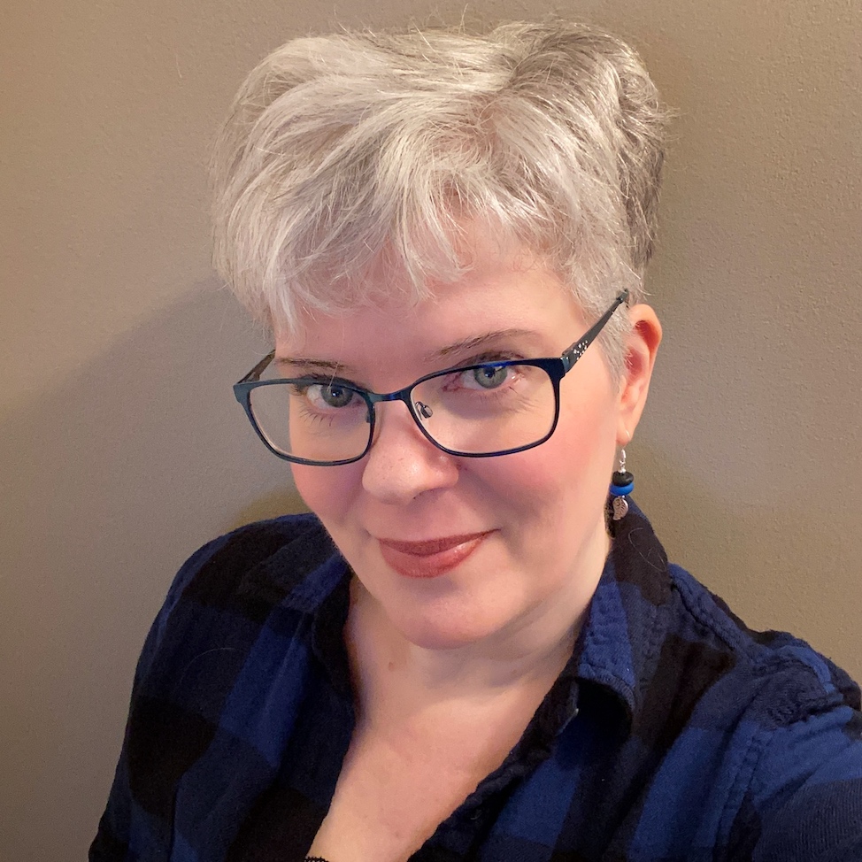 A white woman with short, blonde-white hair and blue eyes wearing black framed glasses and a blue and black checked button up. Her earings match her button up. She is smiling with her mouth closed and taking the photo selfie-style.