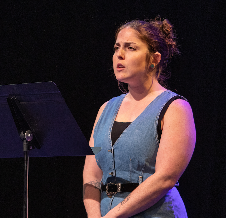 A white woman with dark hair tied back into a bun. She wears a blue dress with a black belt. She is speaking or singing in front of a music stang.