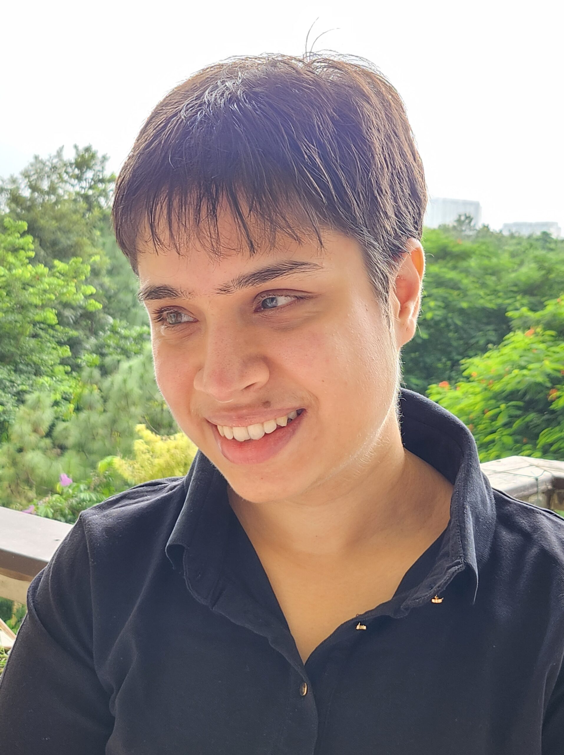 A woman with light brown skin and short, dark hair wearing a dark button up. She is standing in front of trees.