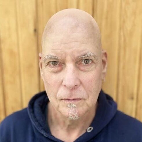 A white man with a shaved head and light brown eyes gazing at the camera. He wears a navy hoodie.