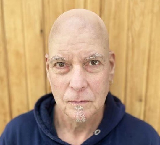 A white man with a shaved head and light brown eyes gazing at the camera. He wears a navy hoodie.