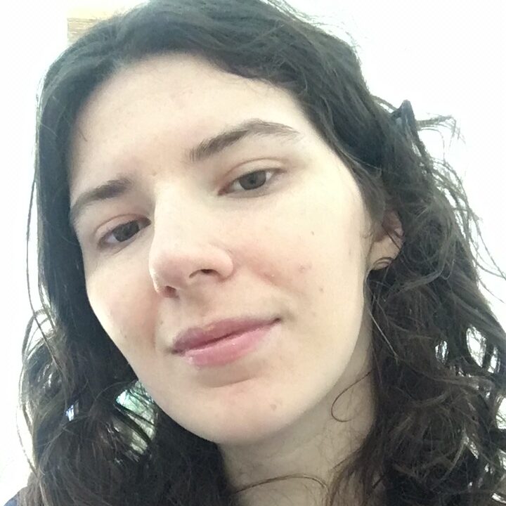 A white person with dark, curly hair smiling at the camera with her mouth closed. They wear a black shirt.