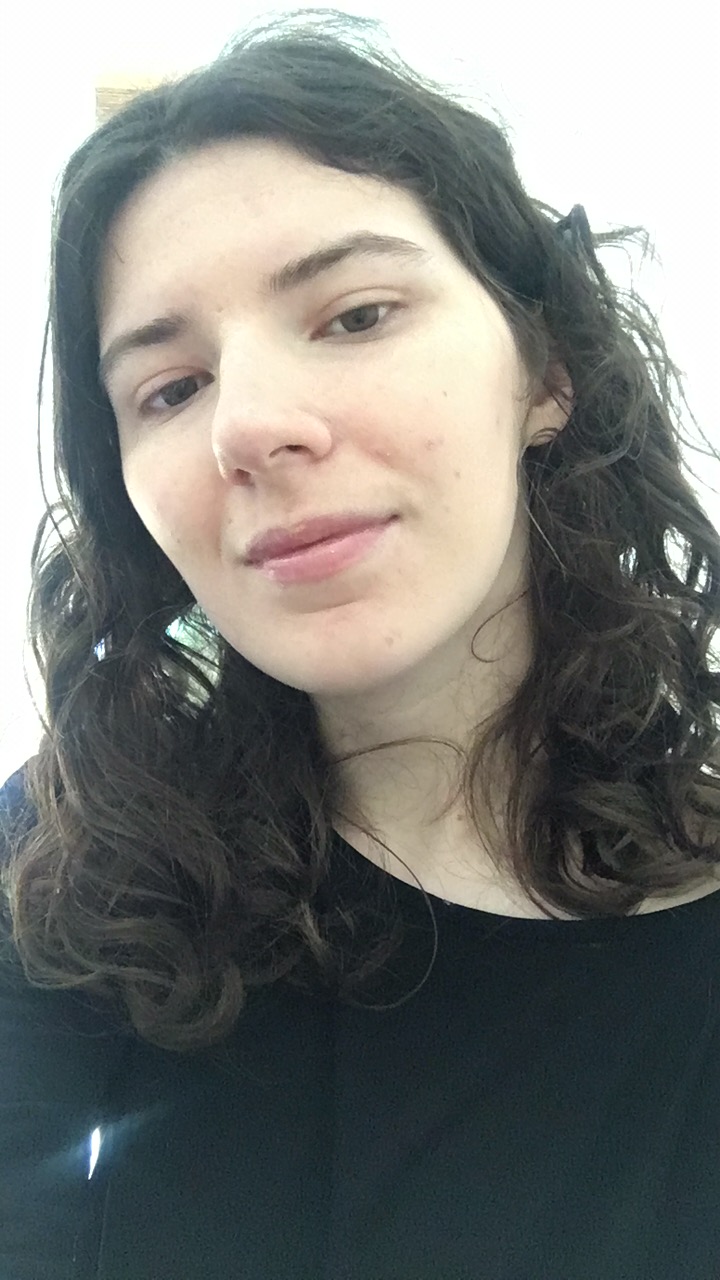 A white person with dark, curly hair smiling at the camera with her mouth closed. They wear a black shirt.